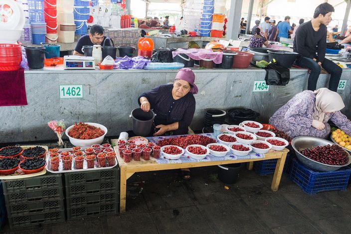 A stallholder in a headscarf sitting behind a trestle table at Chorsu Bazaar banked with bowls of cherries, redcurrants and dark berries, plastic crates stacked at her feet and other traders working along the concrete counter behind her under the market roof.