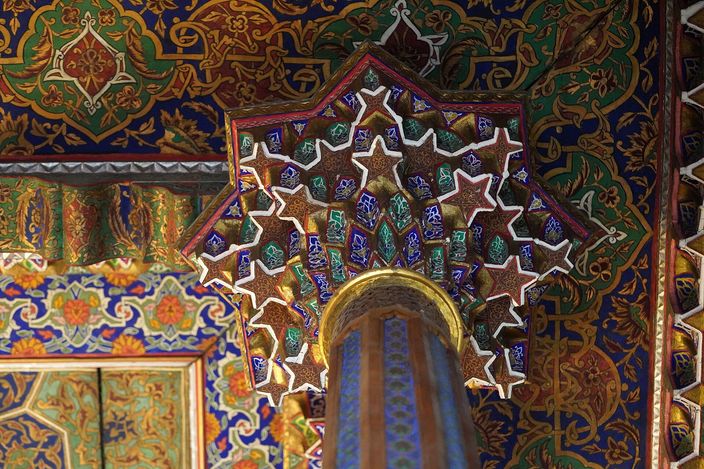 A muqarnas bracket of carved and painted gypsum above a slim blue-and-gold column inside the State Museum of Applied Arts in Tashkent, its interlocking stars outlined in white against green, blue and gilt cells, with densely painted floral panels covering the wall and ceiling around it.