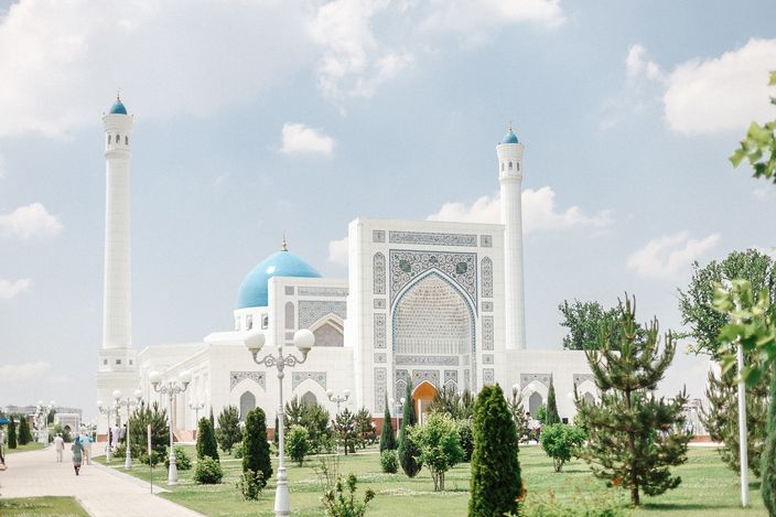 The Minor Mosque in Tashkent across its lawns: a white marble prayer hall with a tall carved portal, a turquoise ribbed dome rising behind it and two white minarets with turquoise caps, young conifers, clipped shrubs and lamp posts lining a path in front under a blue sky with broken cloud.