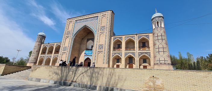 The Kukeldash Madrasah in Tashkent seen from the road below its terrace: a long yellow-brick facade with a tall tiled portal at the centre, two-storey arcaded cells either side, a turquoise dome to the left and a slender minaret to the right, a few people standing on the steps under a clear blue sky.