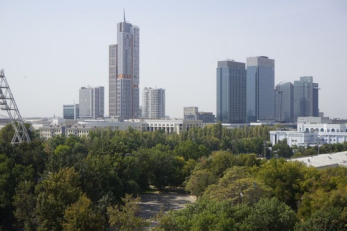 The high-rise towers of the Tashkent City business district rising above a broad green belt of trees, seen from an upper floor, under a hazy sky