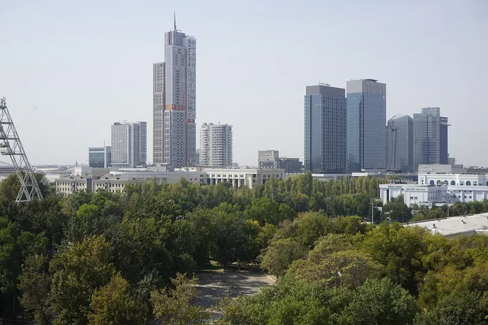 The high-rise towers of the Tashkent City business district rising above a broad green belt of trees, seen from an upper floor, under a hazy sky