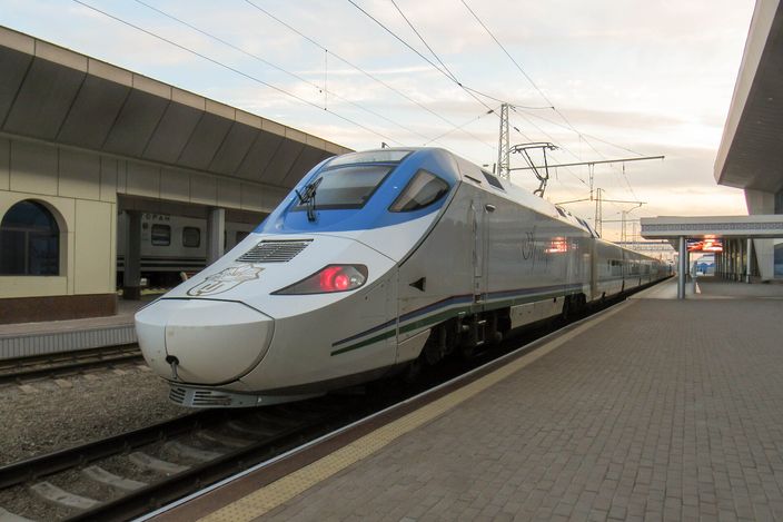 A silver and blue Afrosiyob high-speed train standing at a platform at Tashkent's railway station under a low canopy, its nose towards the camera and the platform empty in the evening light
