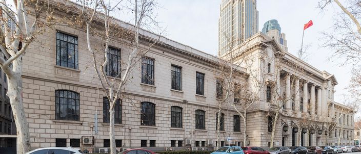 A long grey neo-classical building with a columned portico running down a city street, cars parked nose-to-tail along the kerb, bare plane trees on the pavement and a modern glass tower rising behind it.