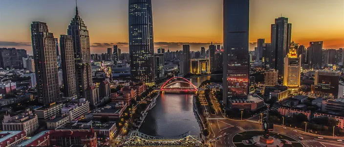 An aerial view at dusk of the Hai River curving between lit high-rise towers, with two illuminated road bridges crossing it and a floodlit roundabout on the right bank.