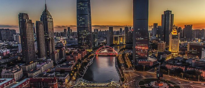 An aerial view at dusk of the Hai River curving between lit high-rise towers, with two illuminated road bridges crossing it and a floodlit roundabout on the right bank.