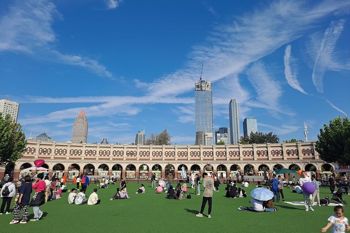 A long arcaded terrace of round arches curving round a stadium, with people sitting and standing on the green turf in front of it and modern glass towers rising behind under a blue sky.