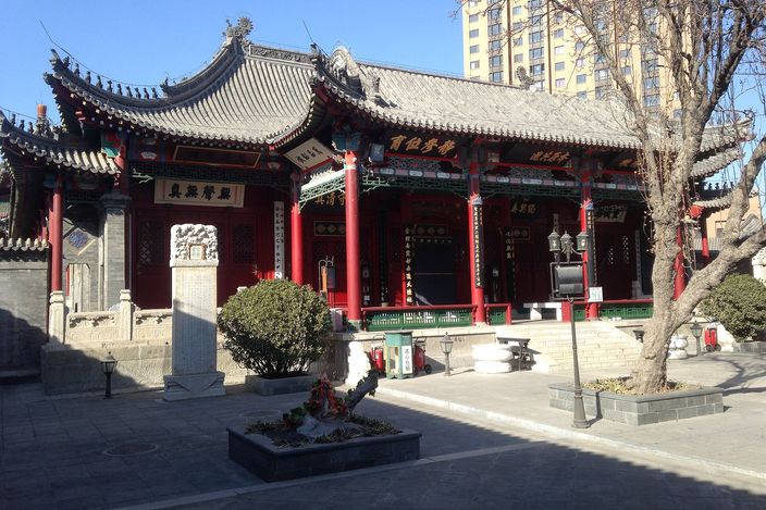 A timber-framed Chinese hall with red columns, carved brackets, gilt-lettered plaques and a grey pitched tiled roof, standing across a swept stone courtyard with a bare tree and a modern apartment tower behind it.