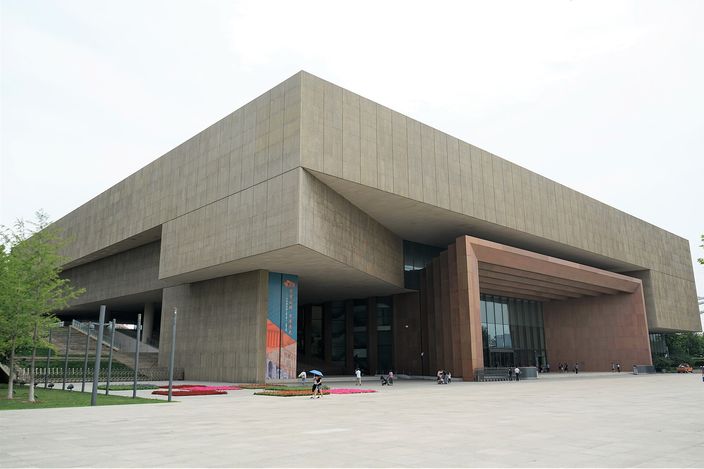A large modern museum clad in pale sandstone, its upper storey cantilevered far out over a colonnade of tall square piers and a glazed entrance, seen across an empty paved plaza on a sunny day.