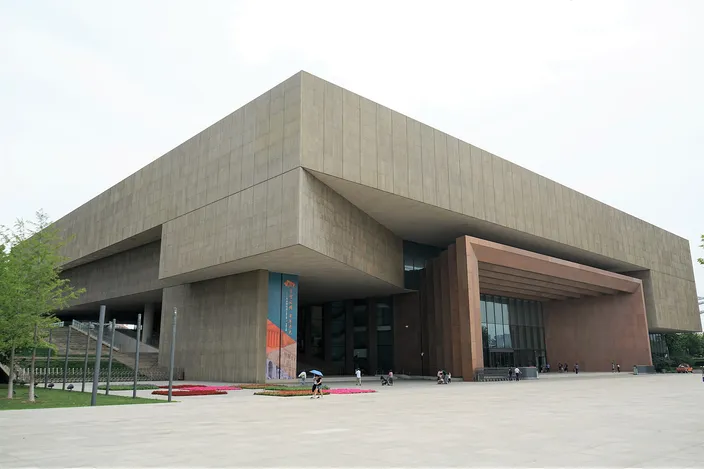 A large modern museum clad in pale sandstone, its upper story cantilevered far out over a colonnade of tall square piers and a glazed entrance, seen across an empty paved plaza on a sunny day.