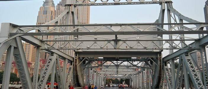 The deck of the Jiefang Bridge seen from one end, framed by its riveted steel lattice towers and cross-girders, with a scooter rider and pedestrians on the near side and tall buildings beyond the far end.