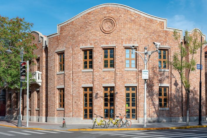 A two-storey brick corner building with an arched gable and tall shuttered windows on a sunlit street in the Five Great Avenues, shared bicycles parked against the kerb and a pedestrian crossing in the foreground.
