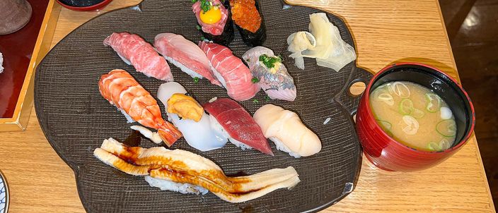 An assortment of Edomae nigiri sushi on a dark plate with a bowl of miso soup, showing pieces of tuna, salmon, egg omelette, simmered sea eel and roe.