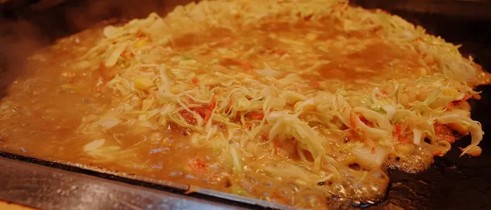 Monjayaki cooking on a flat iron griddle, the loose savory batter spread thin across the hot plate with scattered ingredients and steam rising, in a Tokyo monja restaurant.