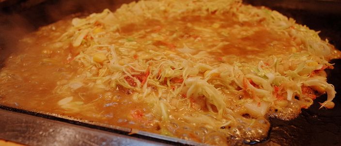 Monjayaki cooking on a flat iron griddle, the loose savoury batter spread thin across the hot plate with scattered ingredients and steam rising, in a Tokyo monja restaurant.