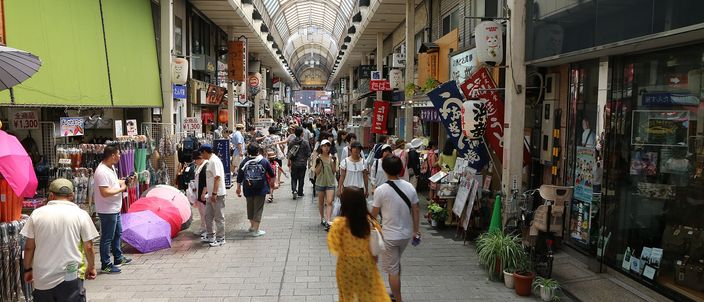 A covered shopping arcade in Asakusa: two rows of small shops under a glazed barrel roof, shoppers walking its length, awnings, umbrellas and goods spilling onto the pavement on both sides