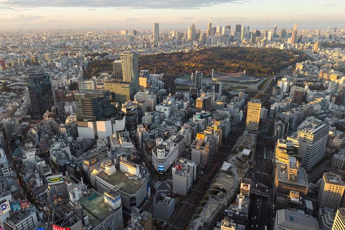 Tokyo at sunset from a high roof above Shibuya: the station district and its blocks immediately below, the dark unbroken green of a large forested park in the middle distance, and the towers of Shinjuku on the horizon in low golden light.