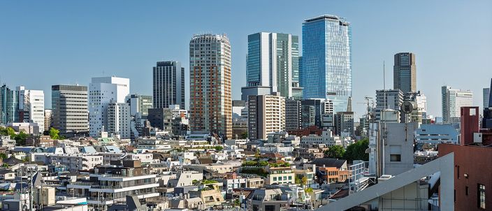 Tokyo on a clear day seen from a rooftop in Harajuku: a dense low city of small buildings and rooftops in the foreground, and behind it a compact cluster of glass and concrete towers rising over Shibuya under a plain blue sky.