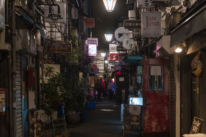 A narrow Golden Gai alley at night, two-storey bar fronts pressed together on both sides under hand-painted signs and a single lamp