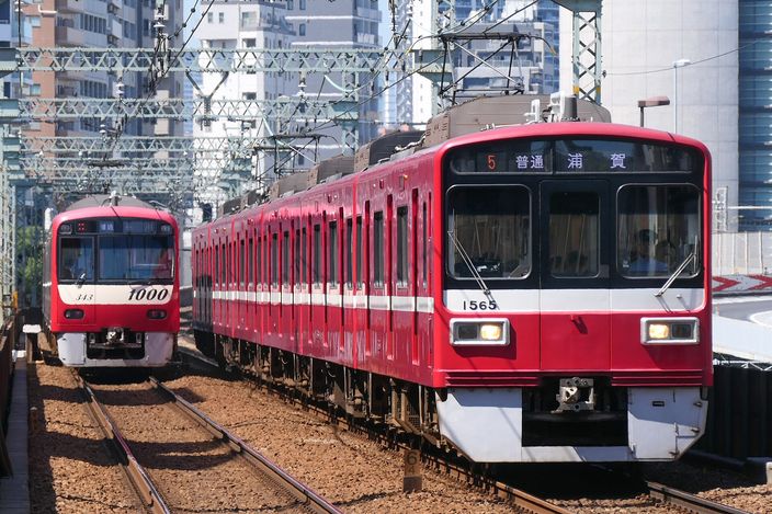 Two red-and-white Keikyu commuter trains on adjacent tracks in a Tokyo cutting, overhead wires above and apartment blocks behind.