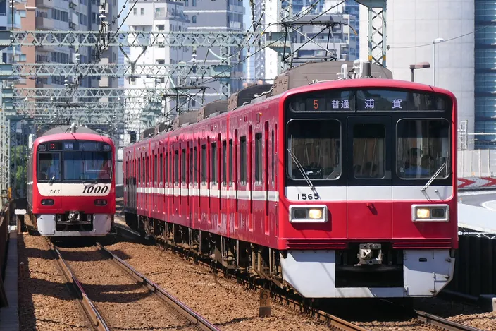Two red-and-white Keikyu commuter trains on adjacent tracks in a Tokyo cutting, overhead wires above and apartment blocks behind.