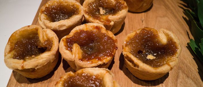 Several round butter tarts on a wooden cutting board, their flaky pastry shells filled with glossy, caramel-dark filling with a crackled top.