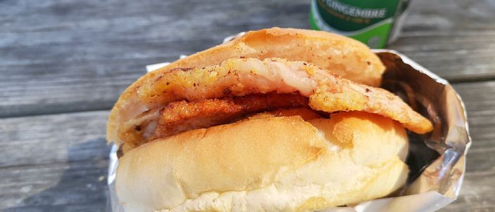 A peameal bacon sandwich: pale cornmeal-crusted back bacon slices on a soft white bun, wrapped in foil on a wooden table.