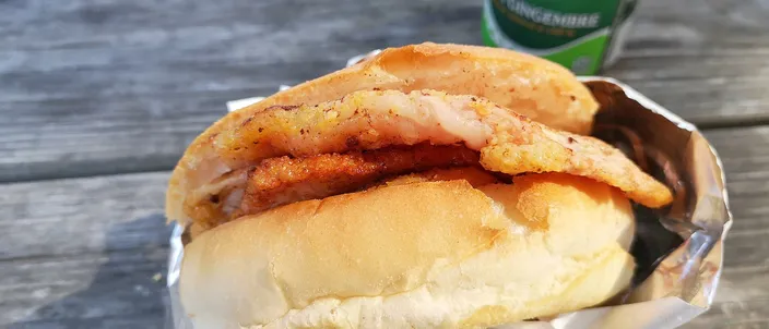 A peameal bacon sandwich: pale cornmeal-crusted back bacon slices on a soft white bun, wrapped in foil on a wooden table.