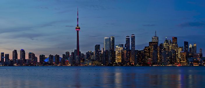 Toronto's downtown skyline at dusk seen across Lake Ontario, the illuminated CN Tower rising among lit financial towers reflected in calm water