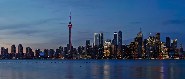 Toronto's downtown skyline at dusk seen across Lake Ontario, the illuminated CN Tower rising among lit financial towers reflected in calm water