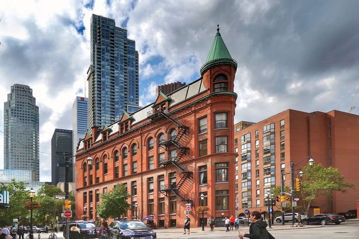 A red-brick Victorian flatiron building on a street corner, its rounded turret capped with a green copper roof, low nineteenth-century blocks beside it and glass towers rising behind under a cloudy sky.