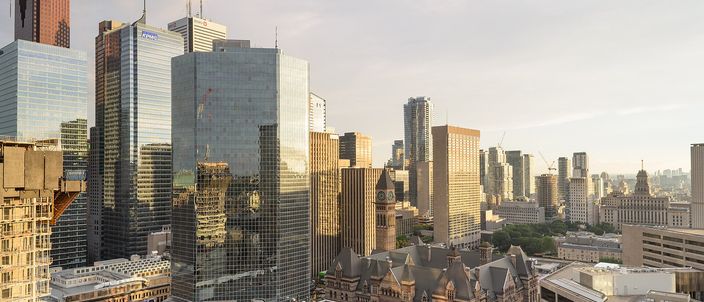 Toronto's downtown financial towers seen from above in low golden light, with the stone clock tower and steep Victorian roofs of Old City Hall standing among the glass in the middle distance.