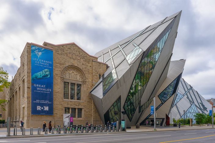 A stack of leaning aluminium-and-glass prisms driven into the side of an older stone museum building, seen from the street corner under a cloudy sky, with a row of bicycles parked along the pavement.