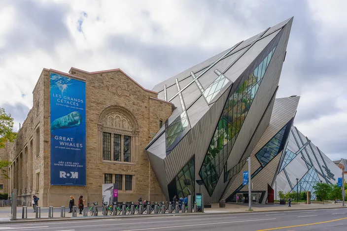A stack of leaning aluminum-and-glass prisms driven into the side of an older stone museum building, seen from the street corner under a cloudy sky, with a row of bicycles parked along the sidewalk.