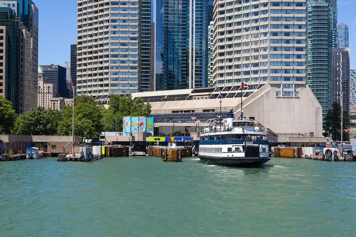 A blue and white passenger ferry crossing open green water towards a low waterfront terminal, with residential and office towers standing directly behind it under a clear sky.