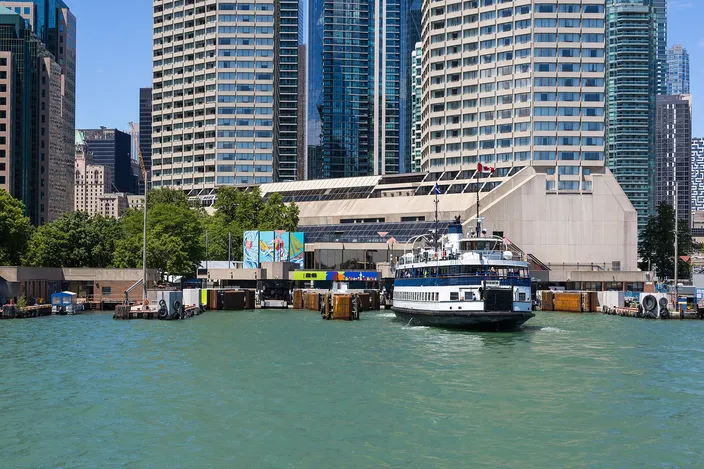 A blue and white passenger ferry crossing open green water towards a low waterfront terminal, with residential and office towers standing directly behind it under a clear sky.