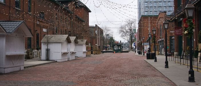 A wide cobbled street between red-brick Victorian industrial buildings, strings of small lights hung overhead, empty white wooden market huts along the left kerb and a blue glass tower rising behind the brick at the far end.