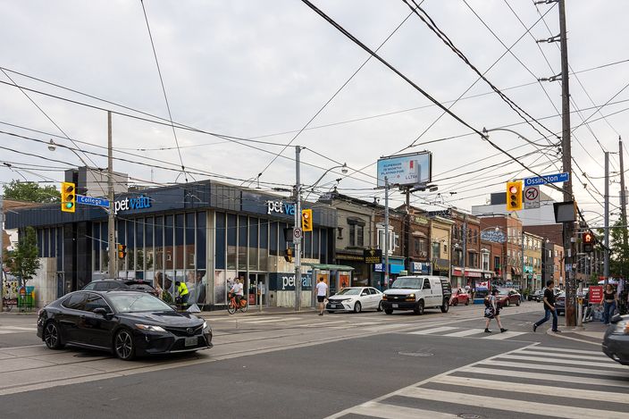 A busy Toronto intersection under a web of streetcar wires, with low commercial buildings, a pet-supply store, pedestrians on a crosswalk and cars waiting at the lights.