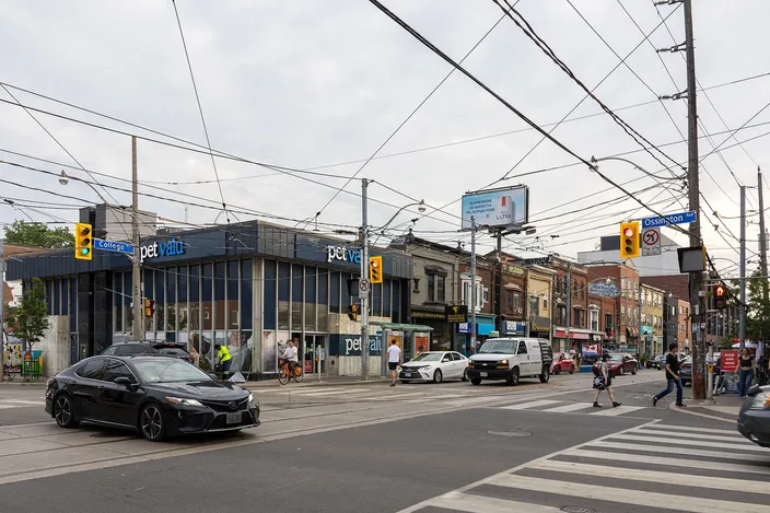 A busy Toronto intersection under a web of streetcar wires, with low commercial buildings, a pet-supply store, pedestrians on a crosswalk and cars waiting at the lights.