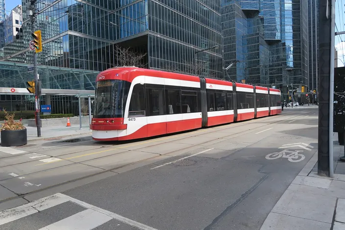 A red-and-white articulated TTC streetcar on tracks crossing a downtown Toronto street, glass office towers behind and a painted bicycle lane in the foreground