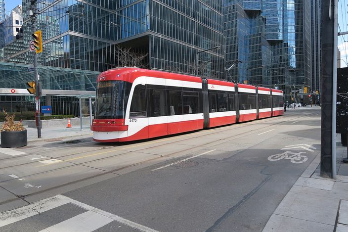 A red-and-white articulated TTC streetcar on tracks crossing a downtown Toronto street, glass office towers behind and a painted bicycle lane in the foreground