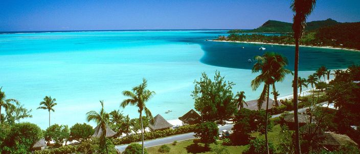 Elevated view over Matira Beach: water grading from pale turquoise near the white sand to deep blue further out, palms and thatched roofs along the curving shoreline