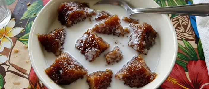 AI-generated image of po'e: glossy caramel-brown pieces of banana pudding standing in a bowl of coconut milk, seen from above, one spoon resting in it, on a floral tablecloth.