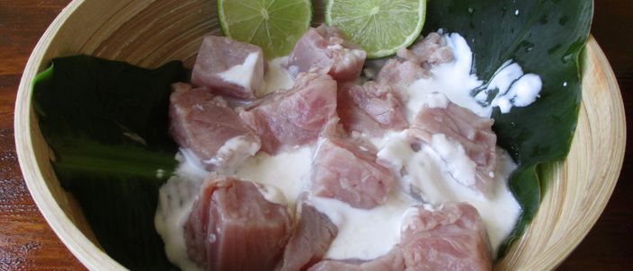 A bowl of poisson cru à la tahitienne — raw tuna cubes bathed in coconut milk, with lime wedges and green leaves as garnish.