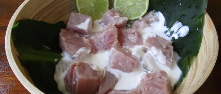 A bowl of poisson cru à la tahitienne — raw tuna cubes bathed in coconut milk, with lime wedges and green leaves as garnish.