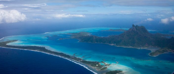Aerial view over Bora Bora's barrier reef and turquoise lagoon toward Mount Otemanu, the eroded volcanic peak at the island's centre, ringed by motu and resort platforms.