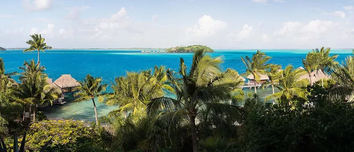 Ground-level panorama over Bora Bora's turquoise lagoon: thatched roofs among palms in the foreground, the water reaching to a low motu on the horizon.