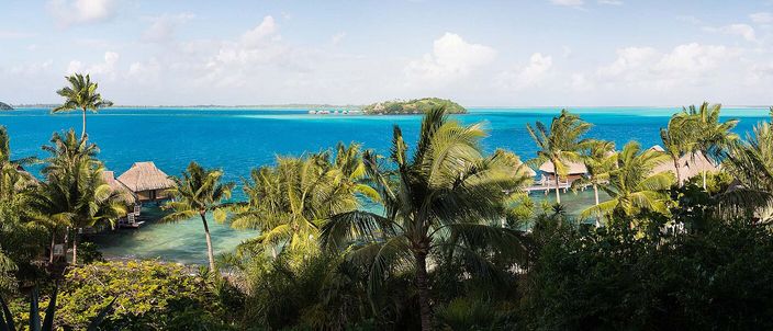 Ground-level panorama over Bora Bora's turquoise lagoon: thatched roofs among palms in the foreground, the water reaching to a low motu on the horizon.