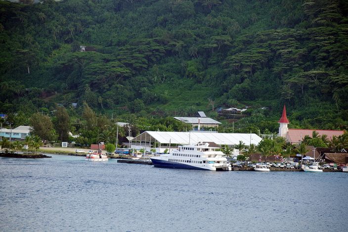 A passenger boat moored at Vaitape's quay, with the village's red-roofed church spire and green hillside behind.