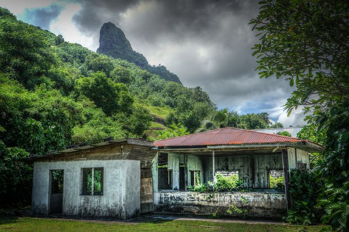 A weathered, open-walled building beside the ring road, with Mount Otemanu's basalt spike looming close under a heavy sky.